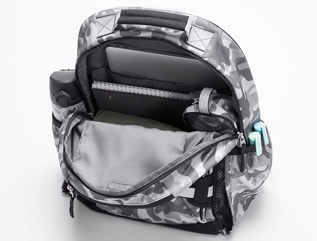 A gray camouflage children’s backpack hangs open, showing school supplies inside