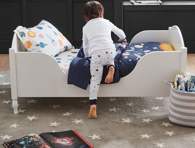 Child in space pajamas climbing on top of shelter toddler bed in space themed montessori bedroom.