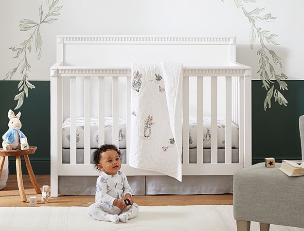 Baby sitting on the floor in front of a crib covered in Peter Rabbit bedding