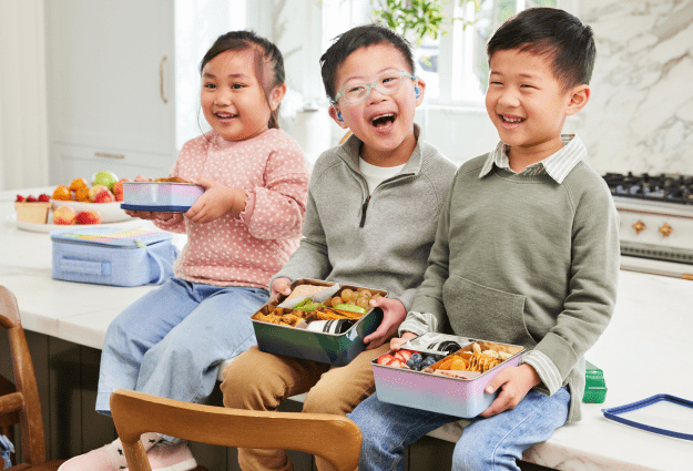 Three kids sit at the kitchen counter holding their stainless steel food containers at lunchtime. 