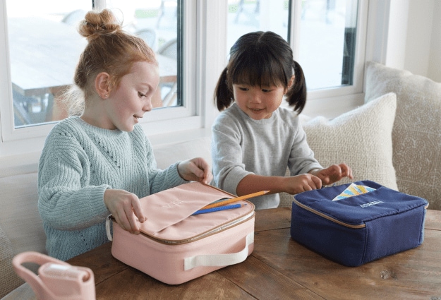 Two girls are sitting for lunch and opening their fun, colorful lunch boxes from Pottery Barn Kids.