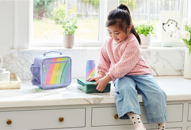 A girl sits atop the kitchen counter closing her food container, readying it to be put inside her Mackenzie lunch bag.