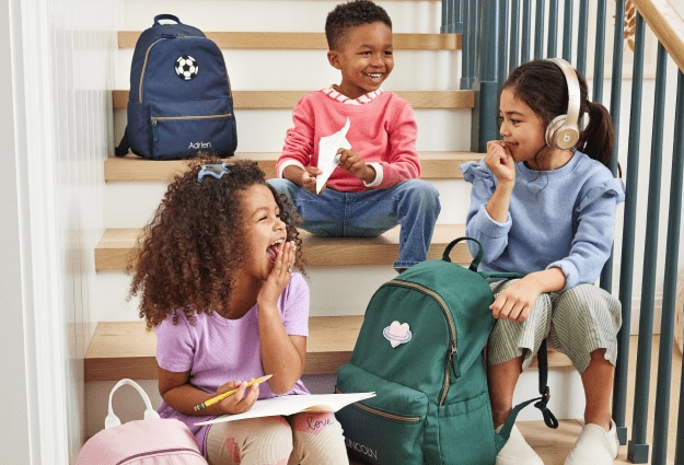 Three kids stand in a stairwell laughing while holding their Colby backpacks from Pottery Barn Kids.