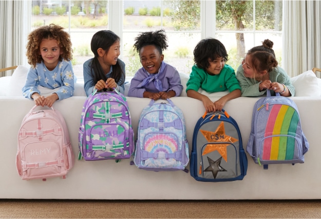 A group of children smile for the camera while proudly posing with their Pottery Barn Kids backpacks and lunch bags.