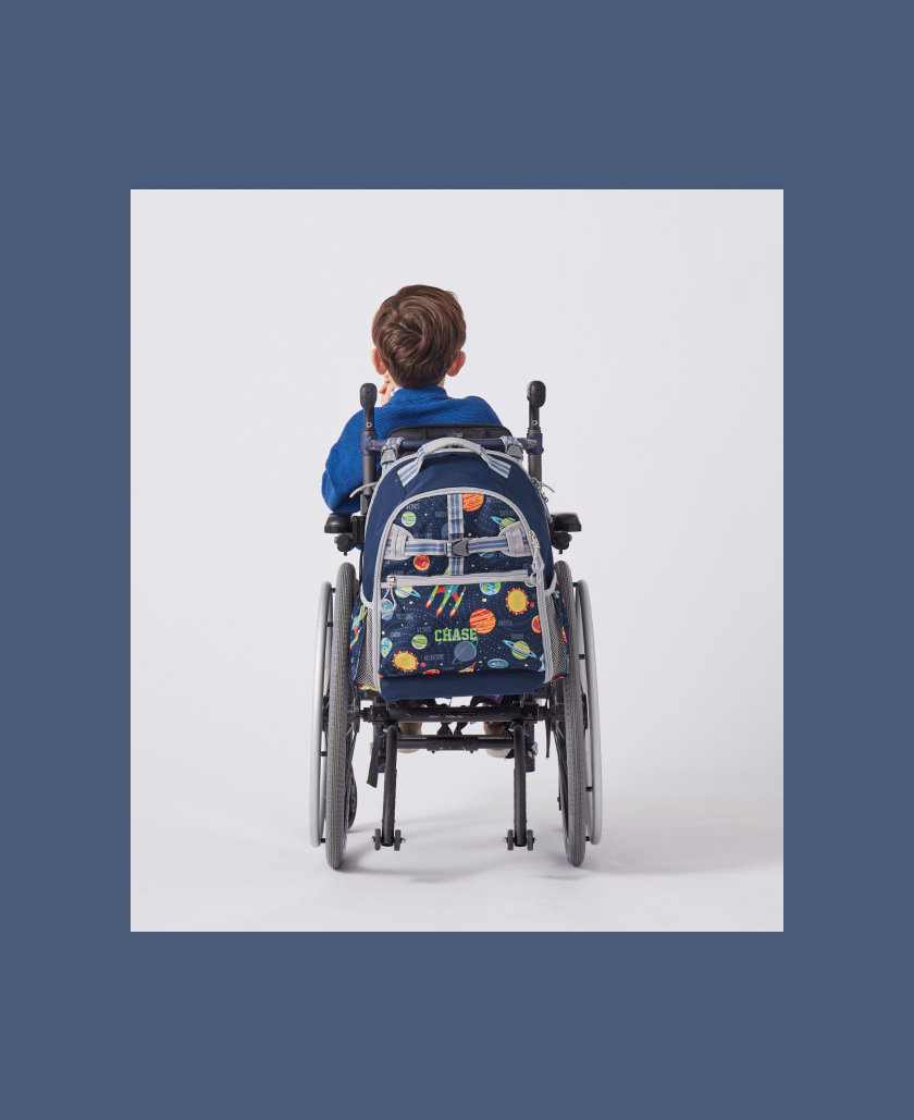 View from behind of young boy in wheelchair with Mackenzie Adaptive Backpack affixed to it.